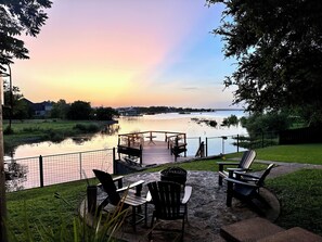 Een terras met Adirondack-stoelen en een tafel, met uitzicht op een rustig wateroppervlak tijdens de zonsondergang.