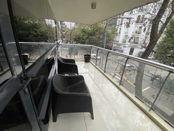 A balcony with black chairs, a glass railing, and a view of a street with buildings and trees.