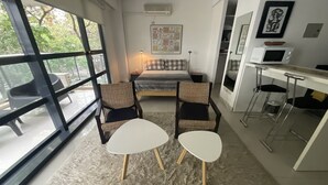 Un soggiorno moderno con una grande finestra, un letto, due poltrone, un tavolino e un angolo cottura con un forno a microonde.