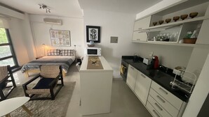 Una camera da letto compatta e moderna con un letto, un comodino e una piccola area scrivania. Adiacente si trova una kitchenette con forno a microonde, fornelli e pensili.