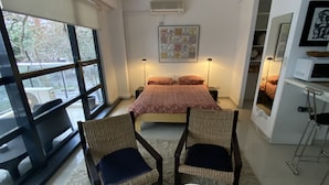 Una camera da letto moderna con una grande finestra, un letto con copripiumino floreale, due poltrone in vimini con cuscini, un tavolino bianco e una ventola a soffitto.