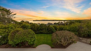 Un jardin aménagé avec un sentier dallé menant à une pelouse, entouré d’une végétation luxuriante et offrant une vue sur une étendue d’eau au coucher du soleil.