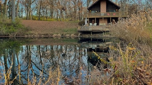 A cabin with a balcony, situated by a pond with reflections, surrounded by trees and overgrown vegetation. - Private Cabin with Pond on Family Farm (Belleville)