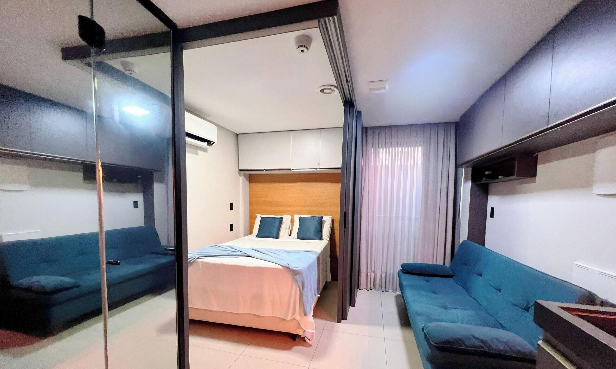 A compact hotel room with a bed, two sofas, a glass partition, and a ceiling-mounted air conditioning unit.