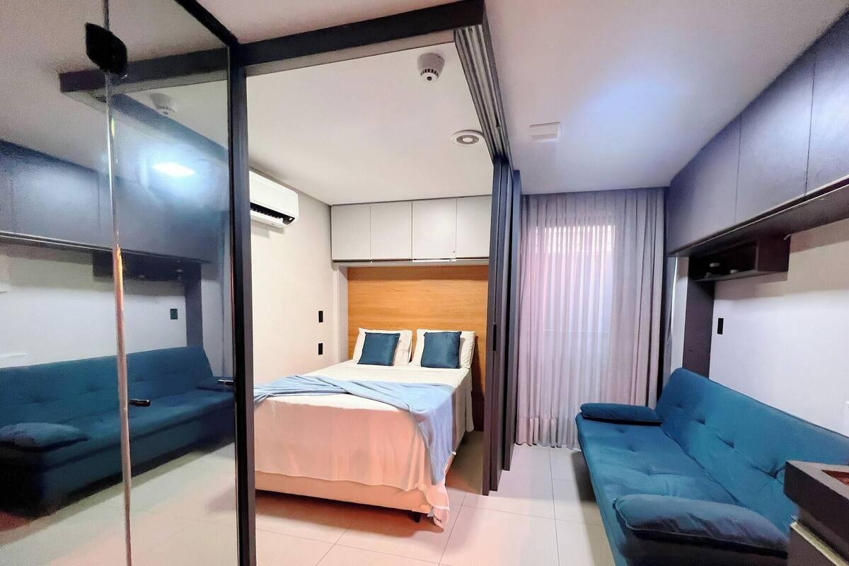 A compact hotel room with a bed, two sofas, a glass partition, and a ceiling-mounted air conditioning unit.