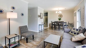 A modern living room with a sofa, coffee table, and a side table with a lamp. There is a dining area with a table and chairs, and a kitchen in the background. - North Charleston Vacation Rental Near Cooper River (North Charleston)
