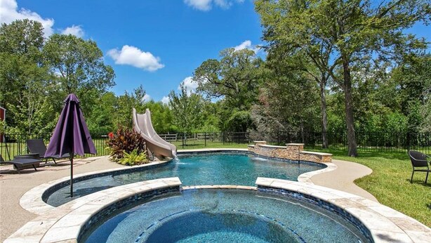 Una piscina con tobogán, rodeada por una terraza de piedra y un área verde con árboles.