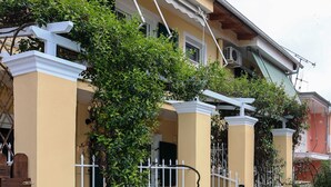 Un edificio residencial con fachada amarilla, barandillas blancas y un pequeño jardín con arbustos y flores.