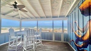 Ett takförsett utomhusområde med en takfläkt, vita Adirondack-stolar och ett bord. Här finns ett stort fönster med utsikt över stranden och havet.
