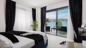 A modern hotel room with a bed, a small table, a chair, and a view of a coastal town. - Beachfront Villa Milka with Pool (Okrug Gornji)
