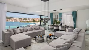 A modern living room with a large sofa set, a dining area, and a view of a coastal town. - Beachfront Villa Milka with Pool (Okrug Gornji)