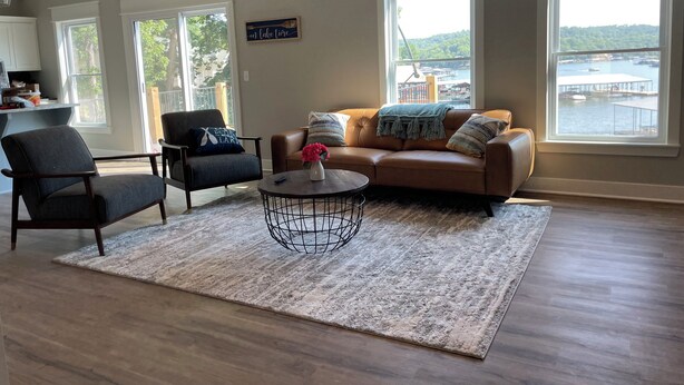 A living room with a brown leather sofa, two armchairs, a coffee table, and a rug. There are large windows with a view of a dock and water. - Lake Escape at Station 46 (Gravois Mills)