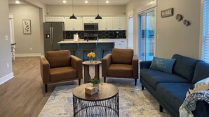 A modern living room with a blue sofa, two brown armchairs, a coffee table, and a kitchen in the background. - Waters Edge at Station 46 (Gravois Mills)