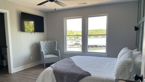 A bedroom with a bed, a chair, a TV, and large windows with a view of a body of water. - Waters Edge at Station 46 (Gravois Mills)
