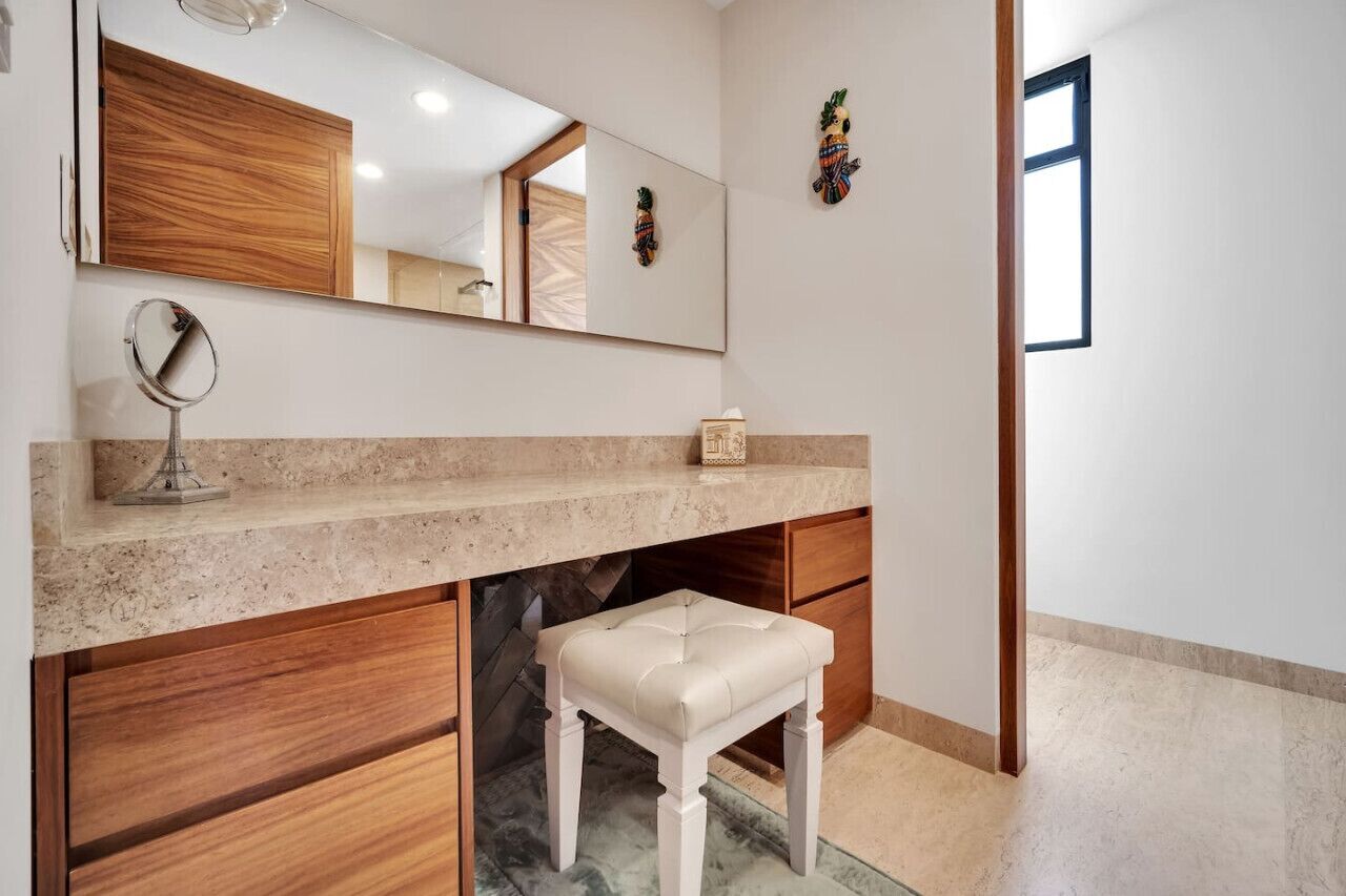 A bathroom with a large mirror, a vanity with a marble countertop, a wooden cabinet, and a white bench.