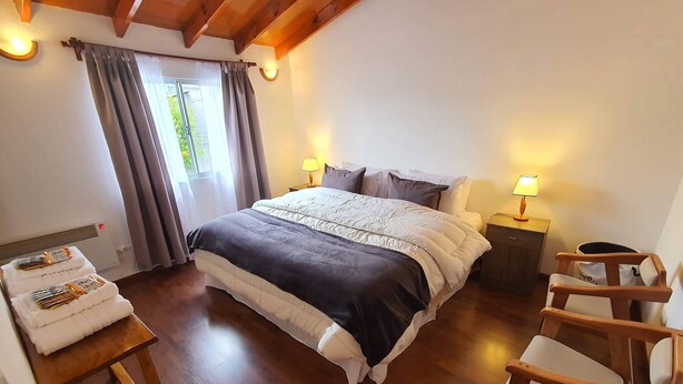 A bedroom with a large bed, two bedside tables with lamps, a wooden chair, a small table with towels, and a window with curtains. - Apartamento Hermoso y muy Cómodo (Ushuaia)