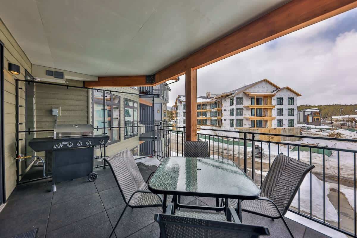 A balcony with a grill, table, and chairs, overlooking a snowy landscape and other buildings.