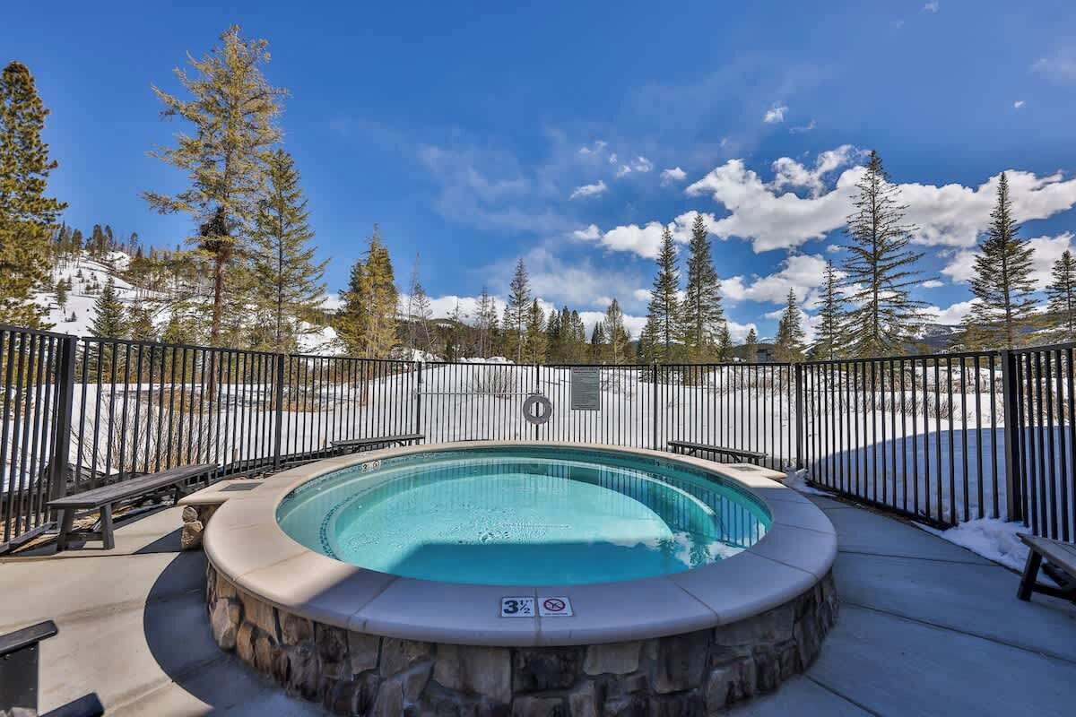 Un jacuzzi rodeado por un muro de piedra y una cerca de metal, frente a un paisaje nevado con árboles y cielo despejado.