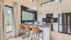 A modern kitchen with a large island, bar stools, stainless steel appliances, and large windows offering a view of the outdoors.