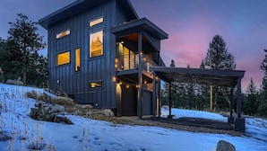 A modern two-story house with a balcony, a covered outdoor area, and a snowy landscape.