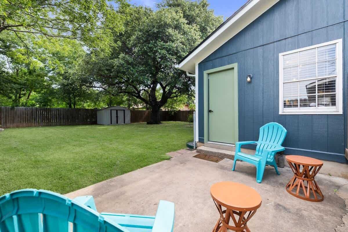 Un patio trasero con una casa azul, césped, una valla de madera y mobiliario exterior, incluyendo dos sillas y dos mesitas.