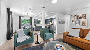 A modern living room with a sofa, armchairs, and a glass coffee table. There is a kitchen in the background with a dining area. - The Oak on 6th Walk to the Historic Square (Georgetown)