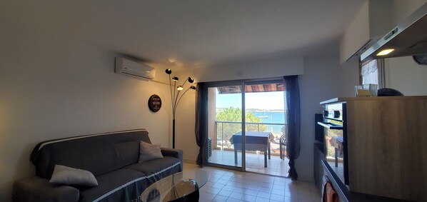 A living room with a sofa, a coffee table, a TV stand, and a view of the sea through a sliding glass door.