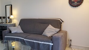 Ein Wohnzimmer mit grauem Sofa, zwei Kissen, einem Glastisch, einer Lampe, einem kleinen Beistelltisch mit Dekoration und einer an der Wand befestigten Ölflasche.