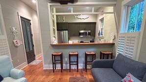 A modern living room with a grey sofa, a patterned ottoman, and a blue couch with a lobster pillow. A wooden coffee table is in the center. The room has wooden flooring and a large window with shutters. - "Nauti-Jayne" coastal cottage in Historic Old Town Bluffton (Bluffton)