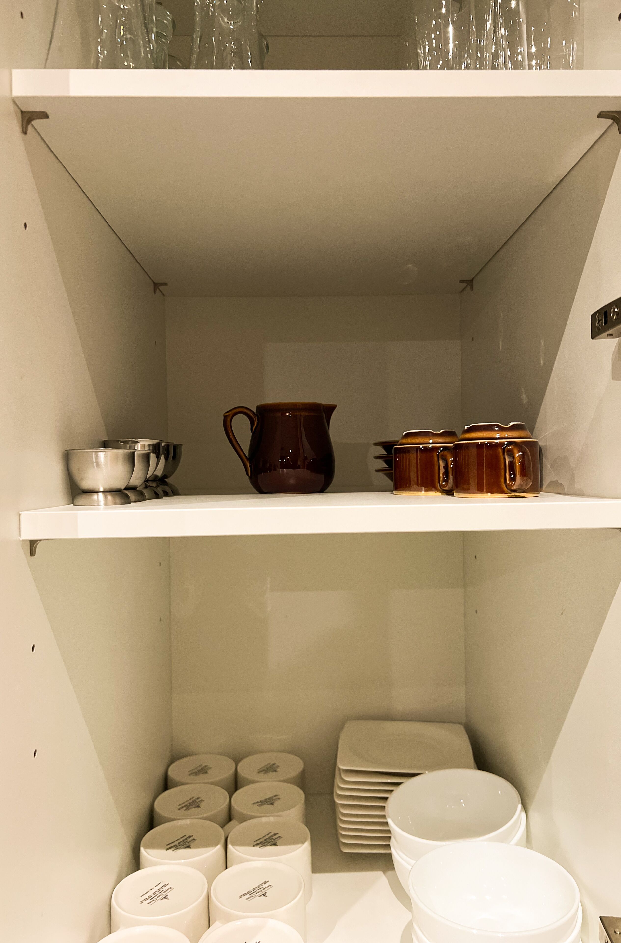 Une cuisine avec des armoires et des étagères blanches, où sont rangés divers ustensiles de cuisine, notamment des bols, un pichet et des assiettes.