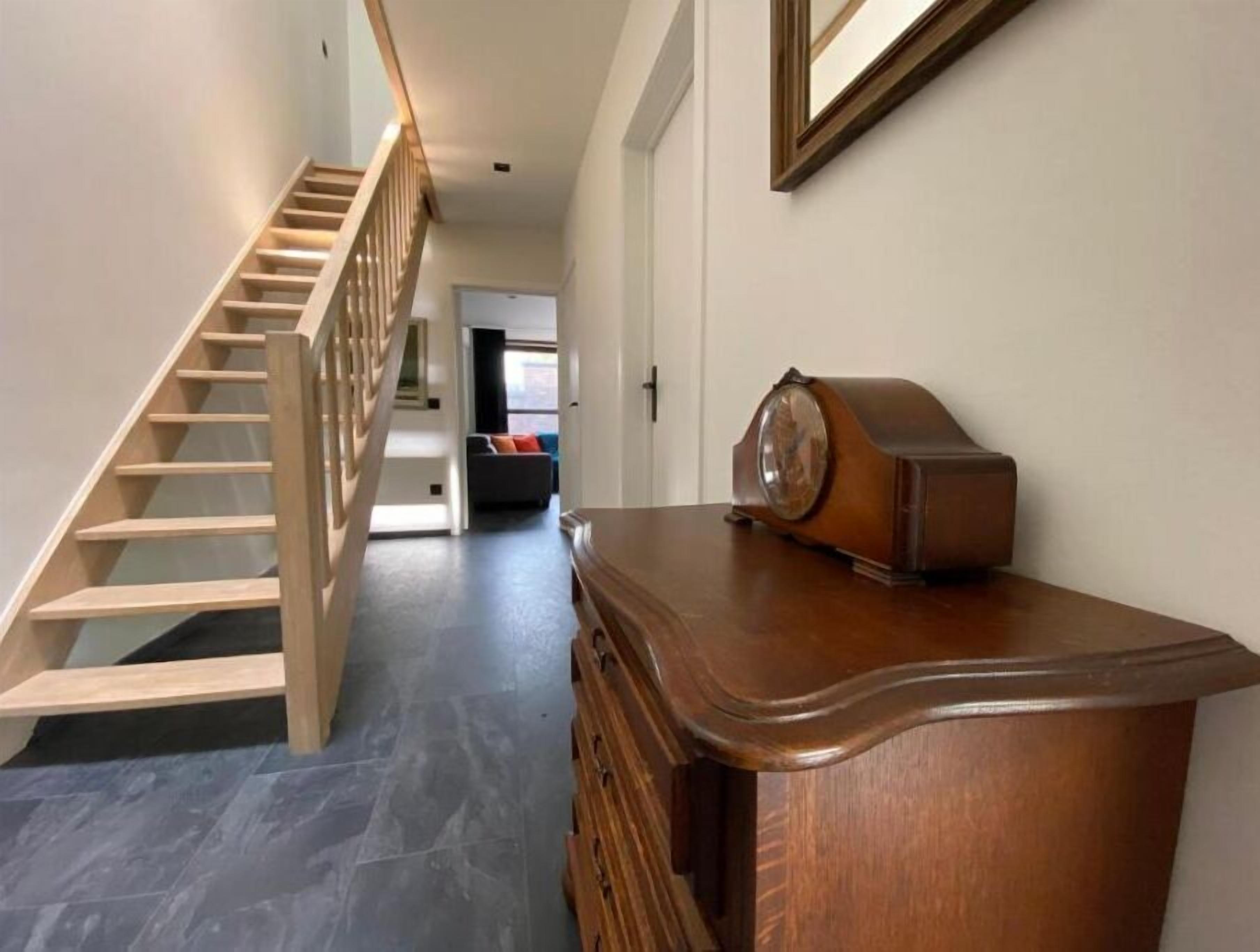 A hallway with a wooden staircase, a wooden dresser with a clock, and a living area in the background.