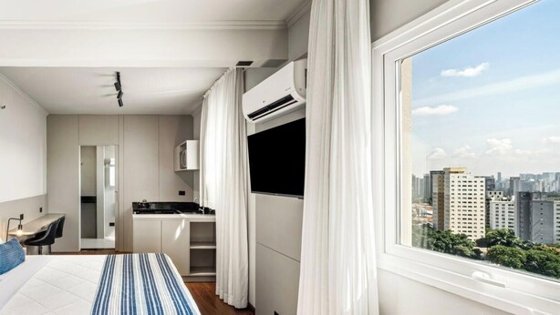 Una habitación de hotel moderna con cama, escritorio, kitchenette y un amplio ventanal con vistas a la ciudad.