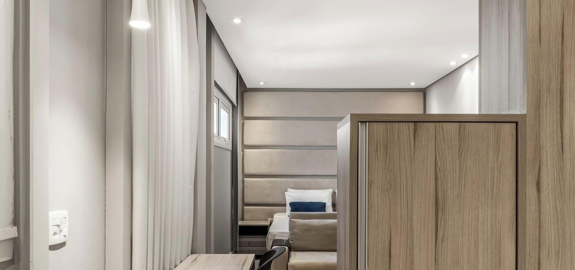 A modern hotel room with a neutral color palette, a bed with a headboard, a seating area with a sofa and chairs, and a wooden partition.