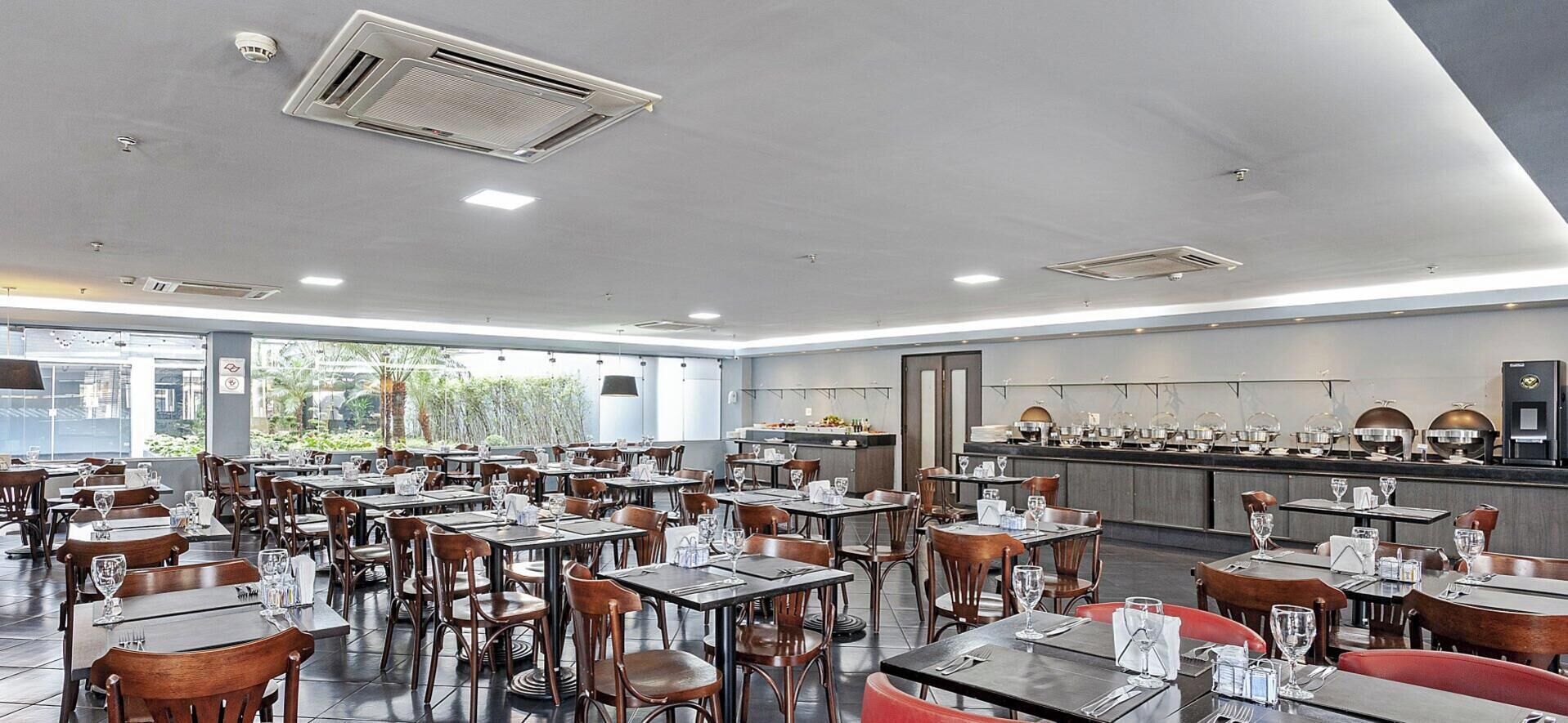 A spacious restaurant with multiple tables and chairs, a kitchen area, and large windows allowing natural light.