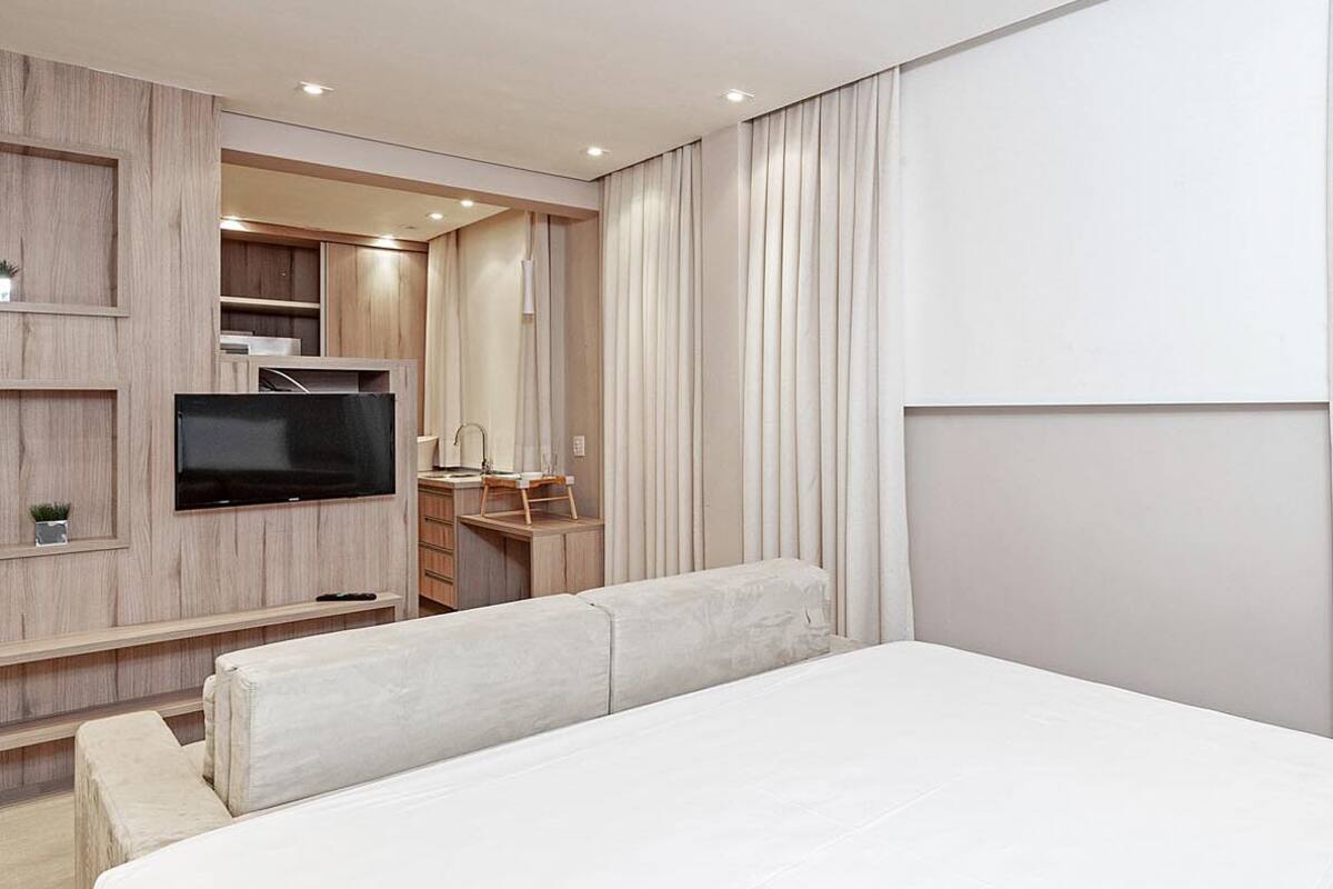 Ein modernes Hotelzimmer mit einem großen Bett, einem flachbildfernseher an der Wand, einem kleinen Schreibtisch mit Stuhl und einem großen Fenster mit Vorhängen.