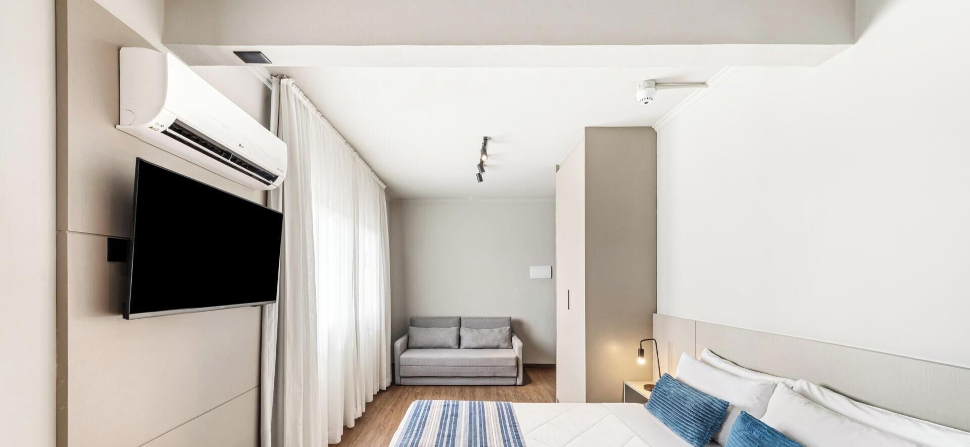 Habitació d'hotel moderna amb televisió de pantalla plana penjada a la paret, un sofà, un llit amb almofadilles i una taula de nit amb una làmpada.
