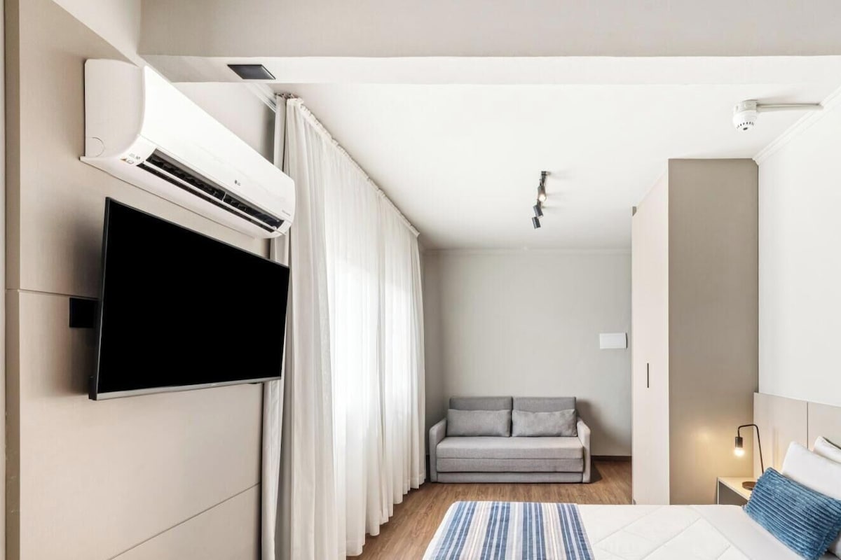 Een moderne hotelkamer met een flatscreen televisie aan de muur, een sofa, een bed met kussens en een nachtkastje met een lamp.