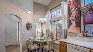 A dining area with a round table, chairs, a chandelier, and a view of the outdoors through large windows. - Garden Patio Home in Chandler, AZ (Chandler)