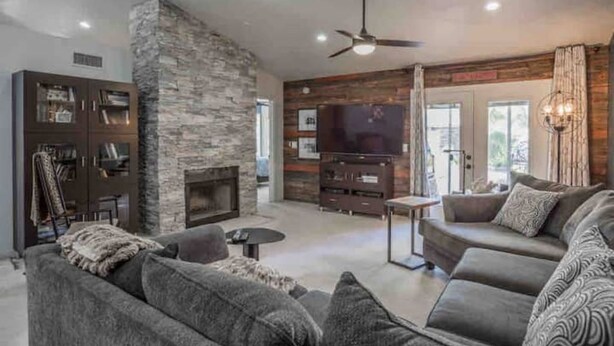 A modern living room with a stone accent wall, a fireplace, a large flat-screen TV, and a cozy seating area with a sofa and armchairs. - Garden Patio Home in Chandler, AZ (Chandler)