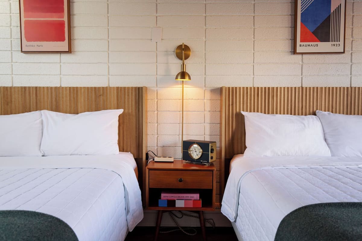 A hotel room with two beds, a nightstand with a lamp, a clock, and a book titled 'Sofia Coppola Archives'.