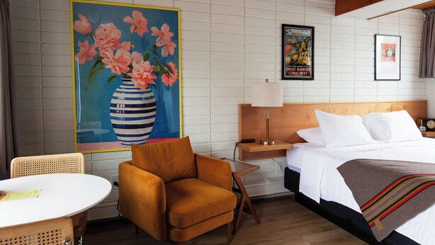 A hotel room with a bed, a nightstand, a lamp, a chair, a table, and a painting on the wall. - The Sierra Motel (Traverse City)