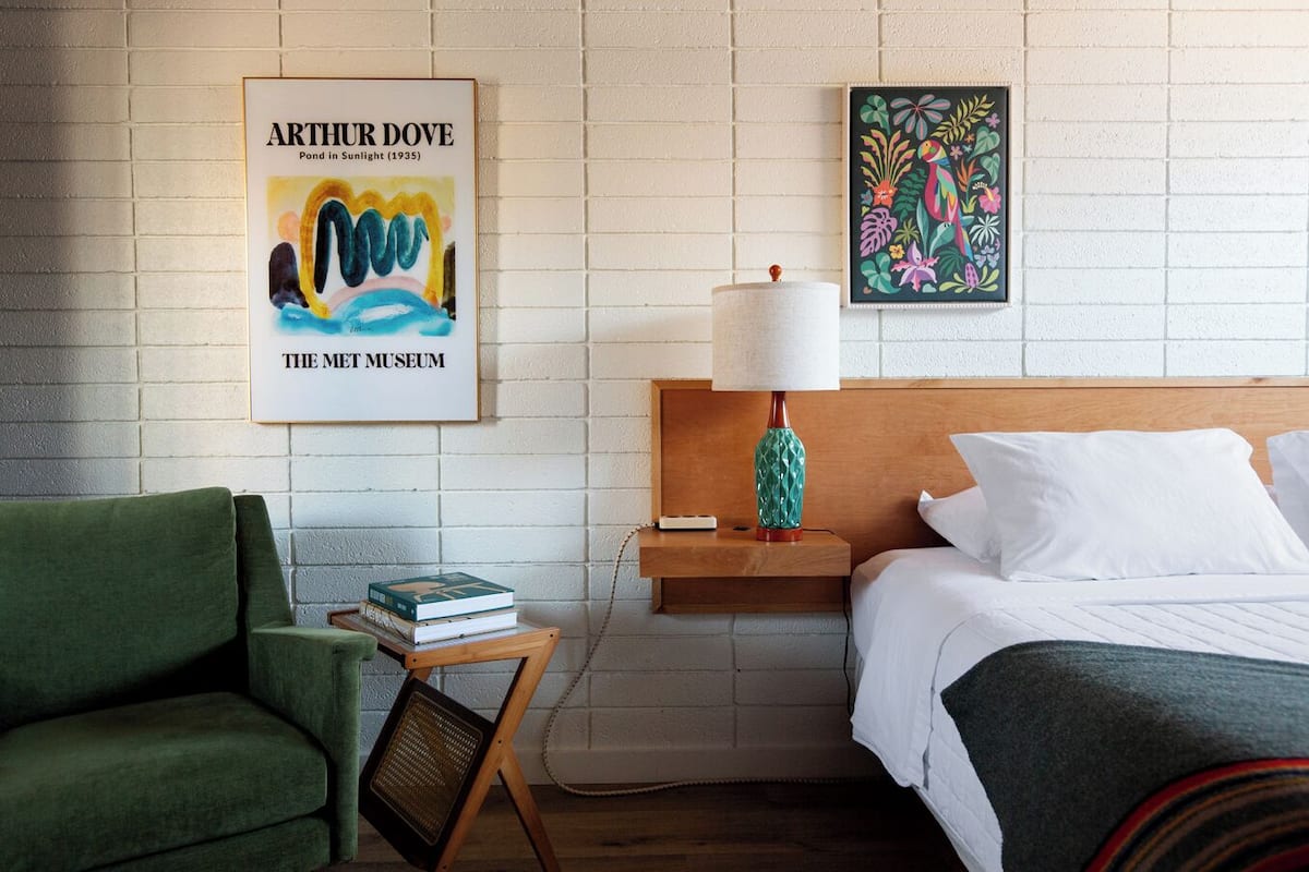 A hotel room with a bed, a green armchair, a nightstand with a lamp, and two artworks on the wall.