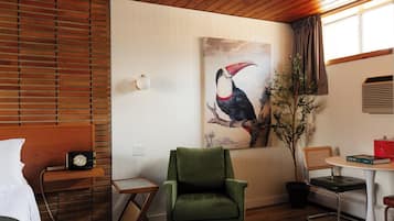 Una habitación con techo de madera, un sillón verde, un cuadro de un tucán, una mesita con un libro y una ventana con cortinas.