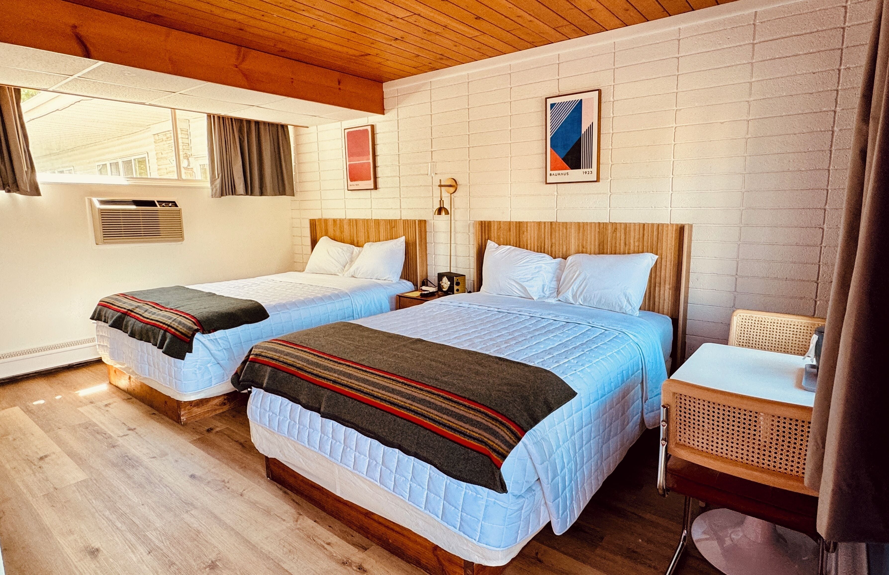 A room with two beds, wooden flooring, a white brick wall, and a small table with a basket.