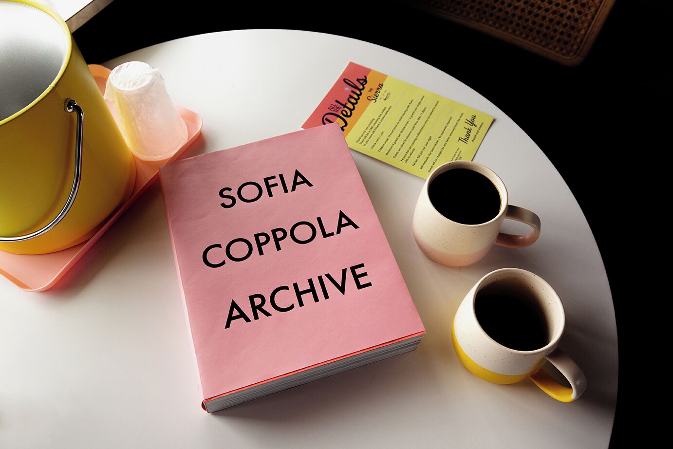 Un livre rose intitulé « ARCHIVES DE SOFIA COPPOLA » est posé sur une table, accompagné de deux tasses à café et d’une théière jaune.