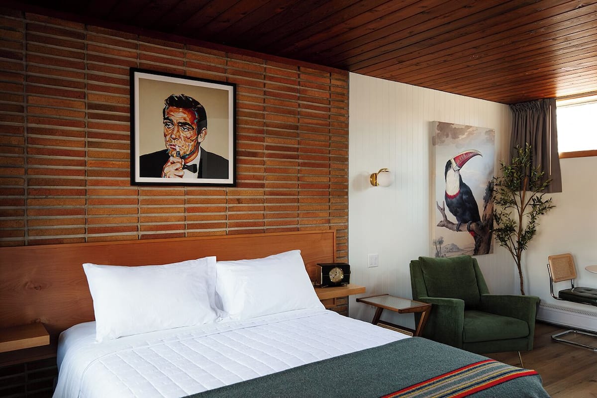 A hotel room with a large bed, a green armchair, a painting of a toucan, and a brick wall.