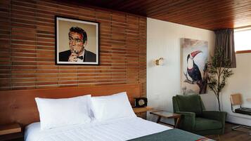 A hotel room with a large bed, a green armchair, a painting of a toucan, and a brick wall.