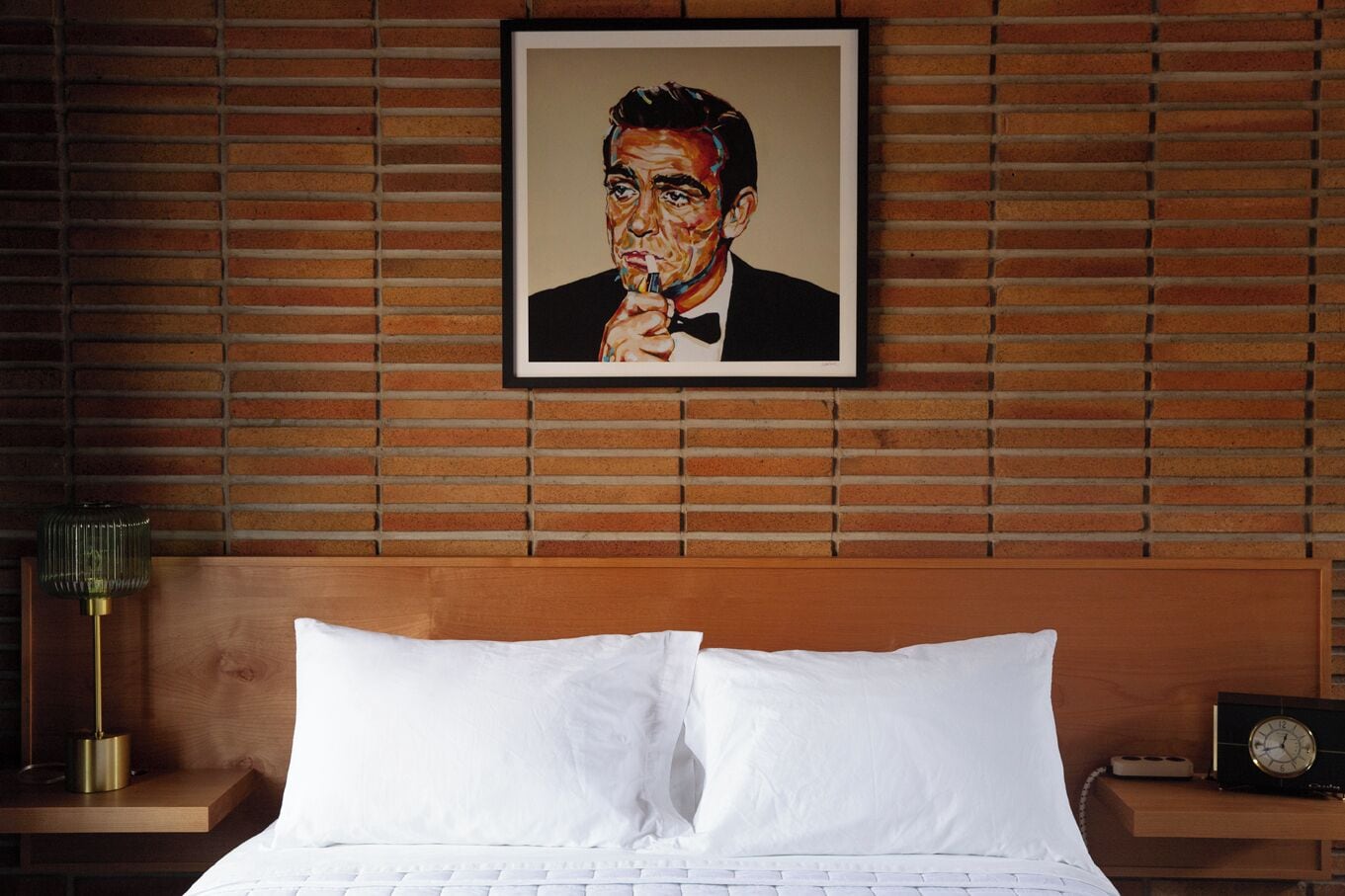 A hotel room with a wooden headboard, a framed portrait on the wall, a bedside table with a lamp, and a clock.