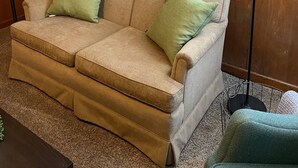 A living room with a beige sofa, two green cushions, a painting of trees, a floor lamp, and a small plant on a side table. - Clean Apartment close to 80/94, I65 Lake Station 2751 DeKalb 1 bed (Lake Station)