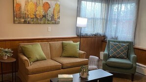 A living room with a brown sofa, a green armchair, a black coffee table, a ceiling fan, and a painting on the wall. - Clean Apartment close to 80/94, I65 Lake Station 2751 DeKalb 1 bed (Lake Station)